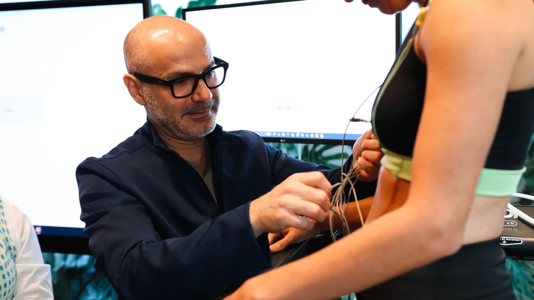 Dr. Mateus conectando eletrodos de eletrocardiograma no torso de paciente em traje esportivo, com monitores LG do laboratório Oxy Recovery ao fundo.
