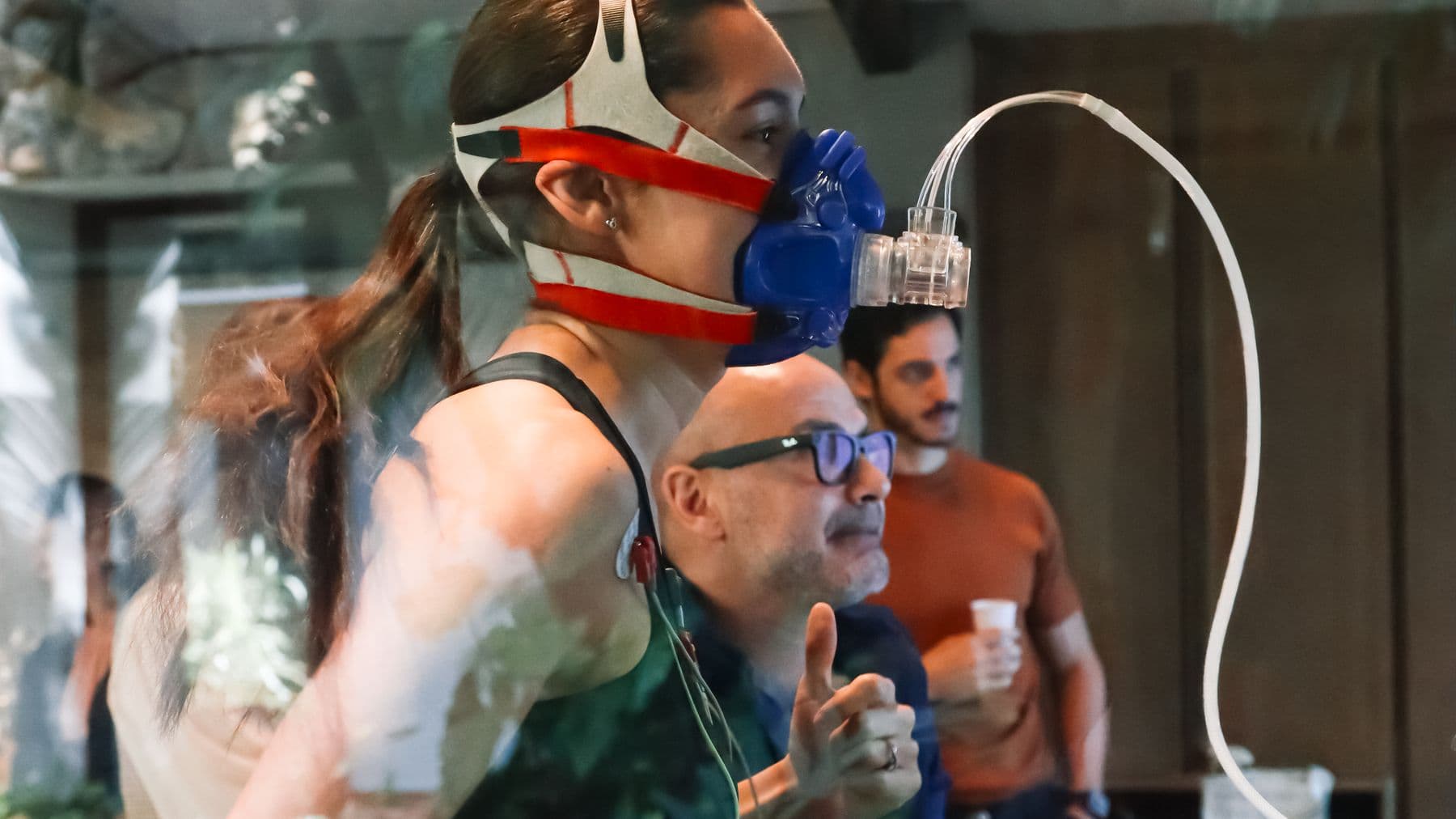 Atleta feminina de perfil em esforço na esteira do laboratório Oxy com máscara de calorimetria conectada ao analisador de gases, Dr. Mateus acompanhando o teste ao lado.