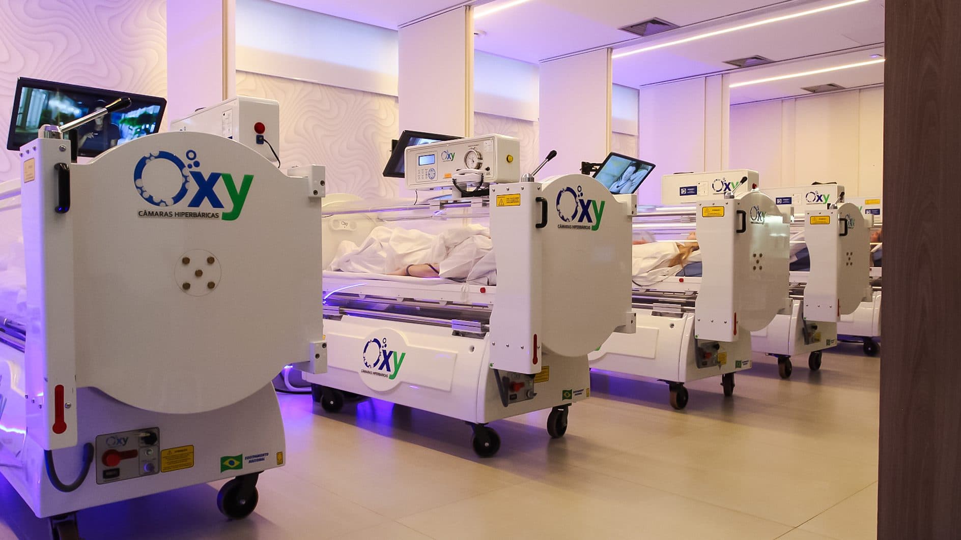 Câmaras hiperbáricas individuais alinhadas em sala clínica com iluminação lilás.